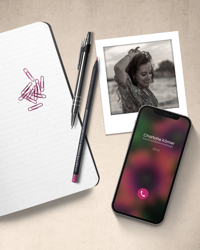 Notizbuch, Stifte, Büroklammern. Ein Handy, auf dem Gerade jmd. anruft. Ein Foto von Charlotte Körner als Polaroid. Die ganze Szene ist von oben aufgenommen worden.